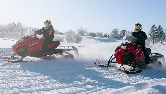 Zwei Personen fahren an einem hellen Wintertag im Deerhurst Resort mit roten Schneemobilen durch den Pulverschnee.