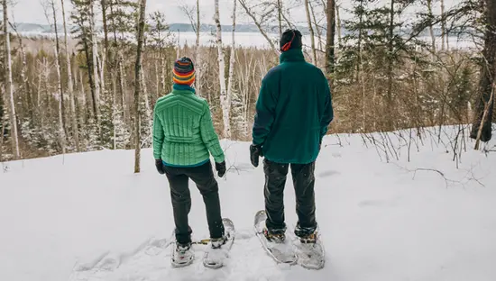Zwei Personen in Schneeschuhen auf einem verschneiten Pfad im Windy Lake Provincial Park in der Nähe von Sudbury und blicken durch hohe Bäume auf einen zugefrorenen See.