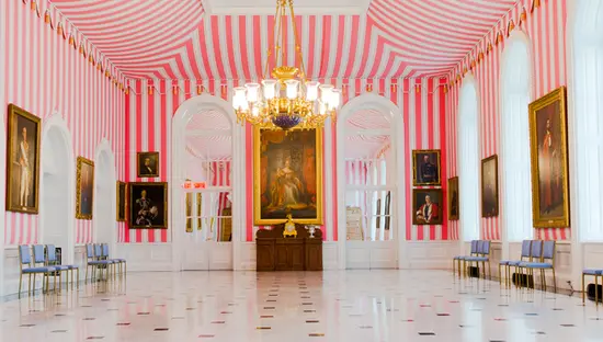  Innenansicht des Zeltraums im Rideau Hall in Ottawa mit kräftig rosa-weiß gestreiften Wänden, goldenen Kronleuchtern und gerahmten Porträts, die den Raum säumen.