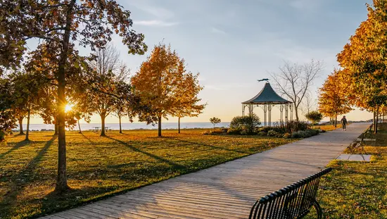  Ein von Bäumen und einer schwarzen Bank gesäumter Holzsteg, der bei Sonnenuntergang zu einem Pavillon an der Uferpromenade von Goderich führt.