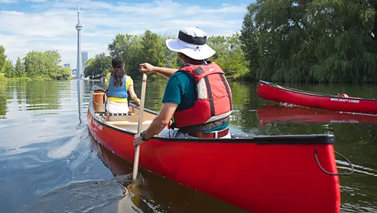 Two people paddling a red canoe towards the CN Tower in Toronto's harbourfront.