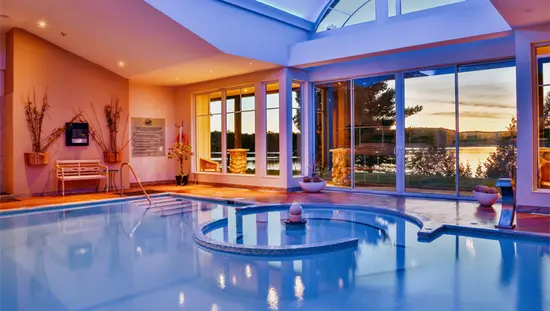 オンタリオ州イーグル レイクにある Sir Sam's Inn & Spa の青い照明、大きな窓、水面に映る夕日の眺めが楽しめるモダンな屋内プール。