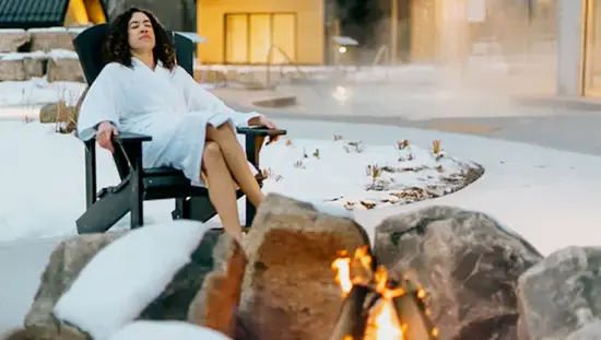 Frau im weißen Bademantel entspannt im Winter auf einem Muskoka-Stuhl im Vettä Nordic Spa, während Dampf aus einem nahe gelegenen Pool aufsteigt.