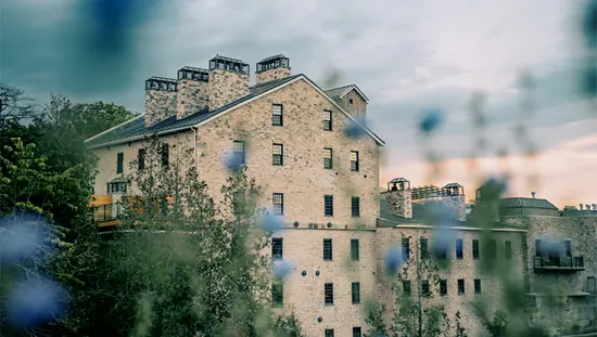 Bâtiment rustique en pierre de l’Elora Mill Hotel & Spa avec ses multiples cheminées, vu à travers des fleurs sauvages d’un bleu tendre au crépuscule.