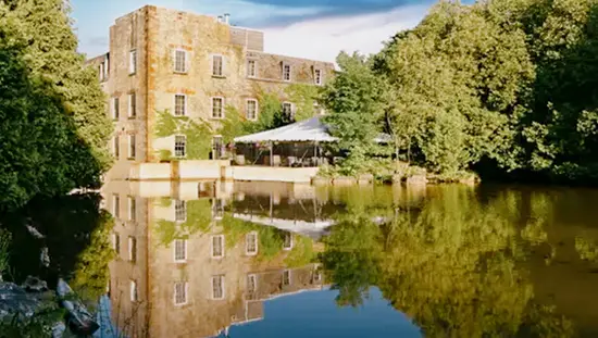 Stone heritage building of Millcroft Inn & Spa reflected in a still pond, surrounded by lush green trees.