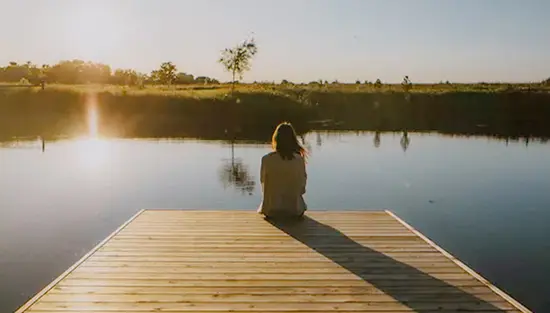 Person sitting quietly at the edge of a wooden dock at Luna Homestead, watching the sun rise over a calm lake in rural Ontario.