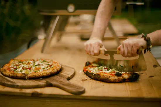 Dos pizzas recién hechas al horno de leña sobre una superficie de madera. Una de ellas se corta con una mezzaluna.