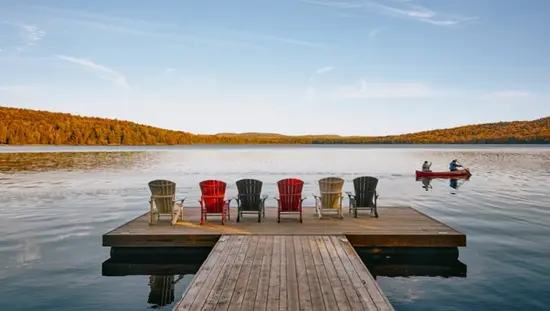 Eine Reihe von sechs Muskoka-Stühlen auf einem Steg blickt auf einen ruhigen See mit Kanus und herbstlichen Farben in der Ferne.