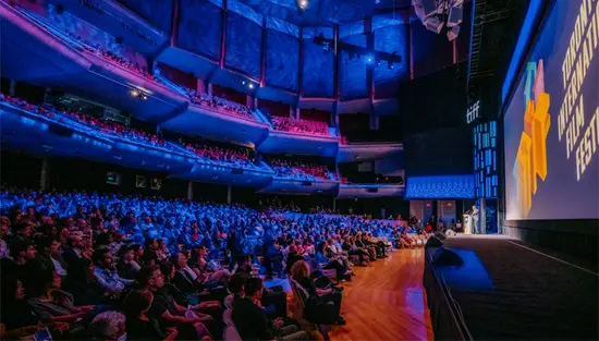 Ein großes, volles TIFF-Theater, das in blaues Licht getaucht ist, und die Teilnehmer blicken auf einen im Scheinwerferlicht stehenden Redner auf der Bühne.