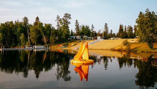 Una playa junto al lago con un tobogán flotante amarillo, canoas y sitios para acampar en el bosque en Cameron’s Beach Campground en Iroquois Falls.