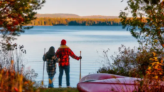 A couple holding hiking sticks looking out onto a still lake surrounded by gorgeous fall foliage.