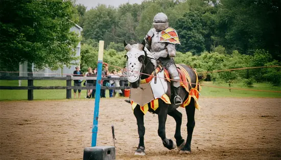A knight riding an armoured horse dressed in red and yellow in front of lush trees and an audience. 