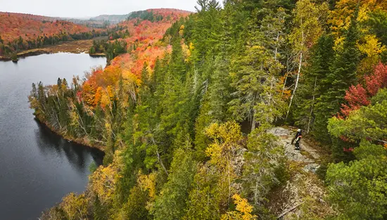 Two hikers stand on a rocky outcrop overlooking a lake below, surrounded by a forest in fall colour.