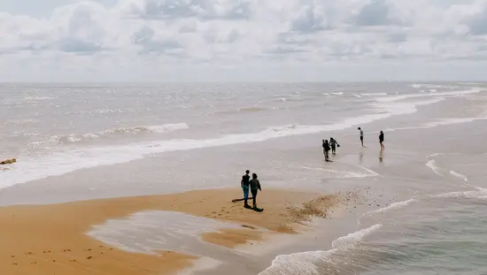 People strolling along a red sandbar stretching into the lake, with gentle waves and clouds on a bright day.