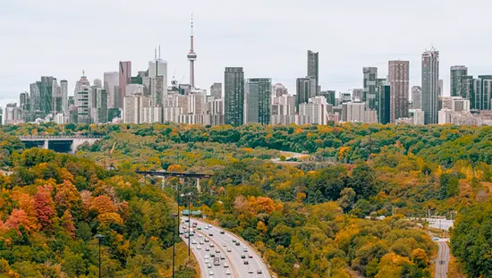 Cityscape view of a highway leading towards the Toronto skyline surrounded by forest with fall foliage.