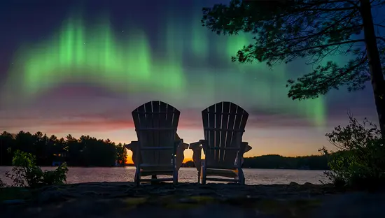Zwei Adirondack-Stühle bieten nachts Blick auf einen friedlichen See in Muskoka, Kanada, und auf die farbenprächtigen Aurora-Lichter, die am Himmel tanzen.