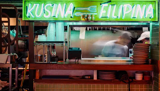 A chef prepares a dish at a food stall with a green neon sign, "Kusina Filipina." Stacks of plates and utensils are in view.