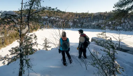 Two people snowshoeing in Northern Ontario stop to admire the view of the forest blanketed in snow across a frozen lake.