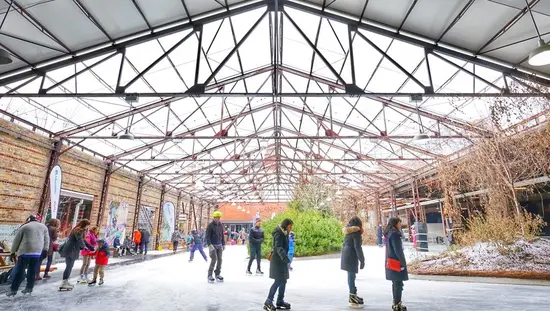 Varias personas disfrutan patinando sobre hielo en una pista de patinaje al aire libre bajo las vigas de la fábrica de ladrillos Evergreen Brickworks.