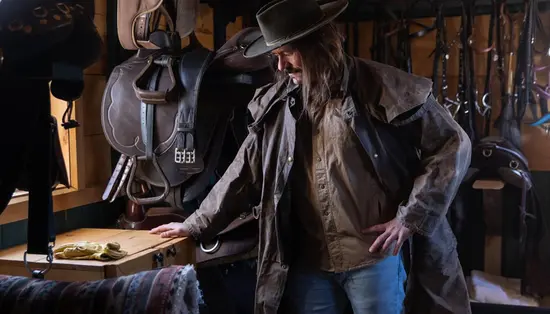A man wearing a cowboy hat and an oilskin duster coat inspects saddles and other horseback riding gear.
