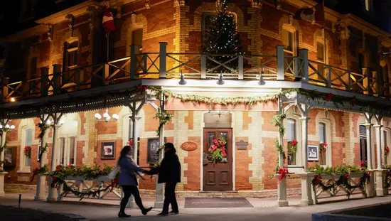 Una pareja tomada de la mano se encuentra en la puerta principal de una posada boutique en un edificio patrimonial decorado con coronas y moños para la temporada navideña.