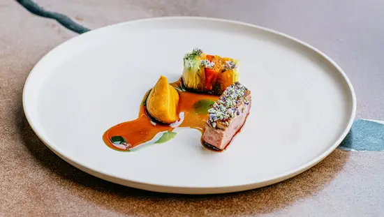 Eloquently served dish of meat and vegetables covered in sauce, with a woman's hand holding a wine glass beside it. 
