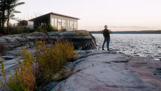 A person standing on a rocky shoreline next to a small cabin overlooking the lake in Northwest Ontario.