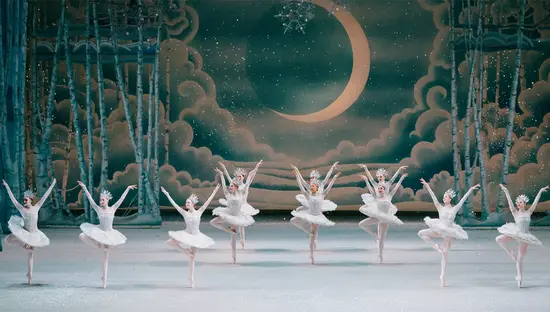 Zwölf Balletttänzerinnen tanzen in einer Reihe mit erhobenen Armen. Im Bühnenhintergrund sind Wolken und ein heller goldener Mond zu sehen.