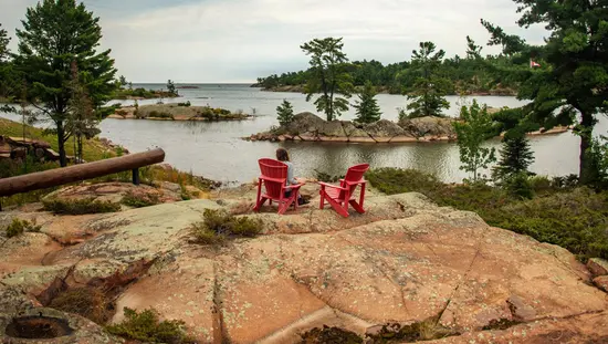 キラーニー近郊のジョージアン湾の海岸線沿いにある象徴的なピンク色の花崗岩の岩の上に置かれた 2 つの赤いマスコーカ チェアのうちの 1 つに座っている女性。