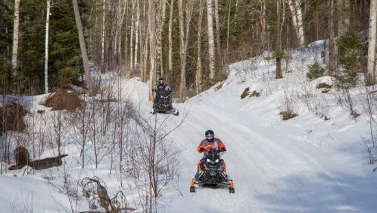 두 명의 스노모빌 운전자가 유명한 라운드 알곤퀸 공원 투어를 따라 숲으로 둘러싸인 눈 덮인 언덕 위로 길을 달리고 있습니다.