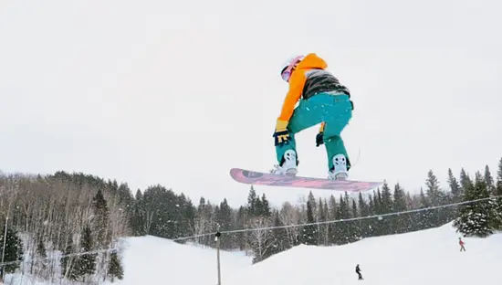 Un snowboarder en el aire realiza un salto en una pendiente nevada, con un bosque a lo lejos bajo un cielo nublado.
