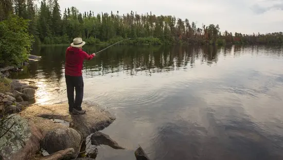A man standing on a rocky shore casts out a fishing line into a calm lake in an Ontario provincial park.