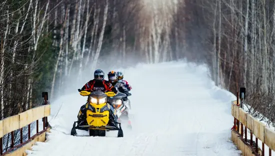 Drei Schneemobilfahrer fahren mit ihren Schlitten über eine schneebedeckte Brücke, im Hintergrund ist ein Wald zu sehen.