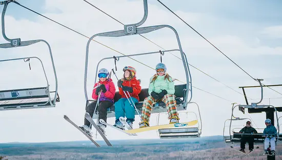 Tres personas viajan en un telesquí con tablas de snowboard y esquís, subiendo por una pendiente nevada en un claro día de invierno.