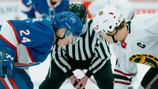2 人のホッケー選手がパックを奪い合っており、1 人は青いジャージを着て、もう 1 人は白いジャージを着ています。審判が 2 人の間にしゃがんでいます。