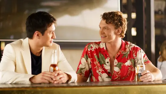 따뜻하고 모던한 분위기의 바에 두 남자가 맥주병을 들고 앉아 담소를 나누며 서로 미소 짓고 있다. 한 남자는 흰색 블레이저를, 다른 남자는 빨간색 꽃무늬 셔츠를 입고 있다.