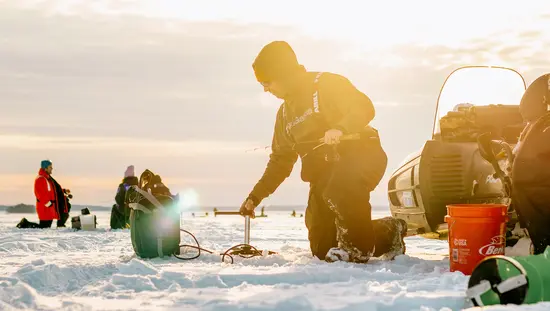 日の出の凍った湖で氷上釣り道具を準備する人々。