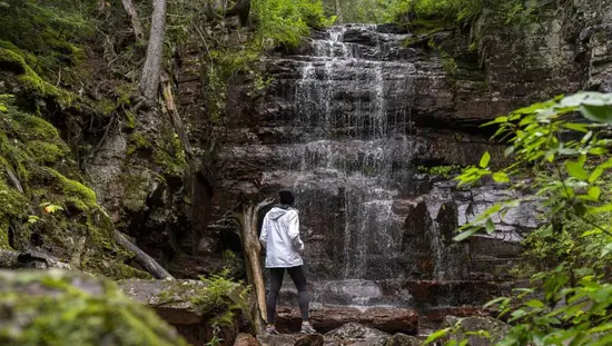 Ein Wanderer hält inne, um einen sanft plätschernden Wasserfall zu bewundern, der von moosbewachsenen Felsen, Bäumen und üppigem Grün im Norden Ontarios umgeben ist.