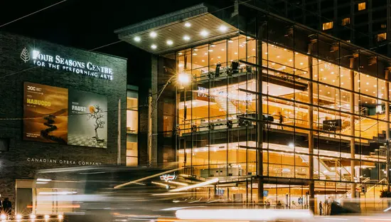 유리로 된 전면을 가진 공연 예술 건물이 번화한 도심 거리에서 밤에 조명을 받아 환하게 빛나고 있다.