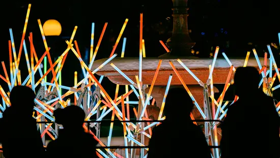 Eine Menschenmenge betrachtet eine nächtliche Lichtinstallation mit bunten, leuchtenden Röhren, die in einem geometrischen Muster angeordnet sind.