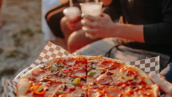 Une pizza fraîchement préparée, des gens tenant des boissons à proximité, sur la plage.