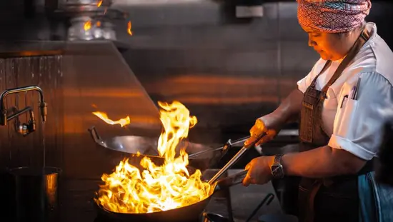 Chef cuisinière faisant habilement flamber un plat dans un grand wok, dans la cuisine du restaurant Kiin de Toronto, étoilé MICHELIN.