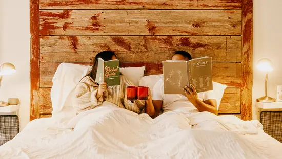 Ein Paar liest im Bett Bücher und hält dabei Tassen in den Händen.