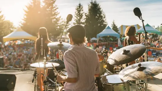 Un asistente al Festival de Música VELD está sentado sobre los hombros de alguien con los brazos extendidos, las luces brillantes del escenario los iluminan.