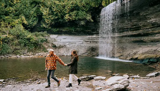 Couple walking hand in hand near a waterfall surrounded by forest.