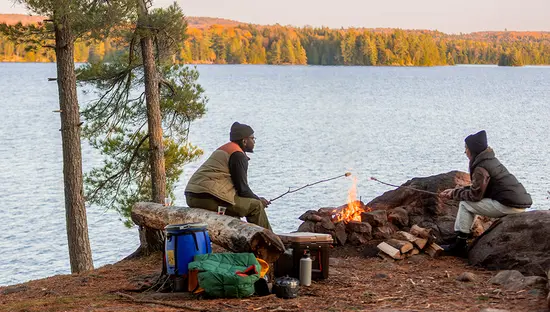 Deux personnes assises près d’un feu de camp sur la rive rocheuse d’un lac faisant griller des guimauves, avec du matériel de camping à proximité.