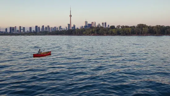 Un homme lance sa ligne de pêche depuis un canoë dans les eaux du port de Toronto, avec la Tour CN et la silhouette de la ville en arrière-plan.
