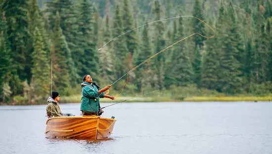 A woman casts a fishing line from a wooden boat on a calm lake surrounded by forest.