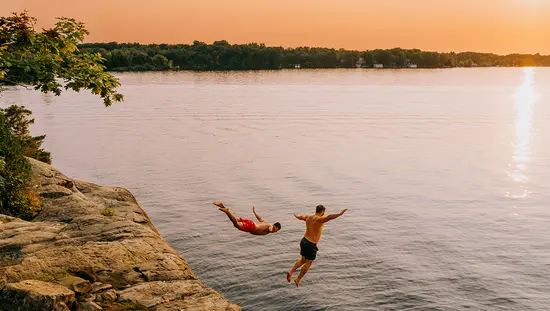 Dos personas saltando desde un acantilado rocoso a un lago al atardecer.