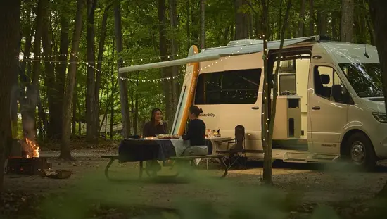 Dos mujeres disfrutan de una cena en una mesa de picnic junto a una caravana estacionada en un camping.